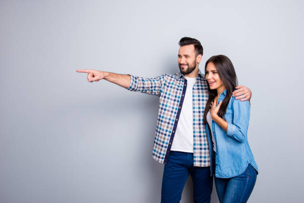 Bearded, smiling man embracing his excited, charming  girlfriend and showing to her something with forefinger over grey background