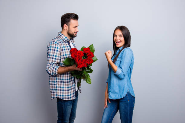 Finally, at last he did it! Man with bristle prepare bouquet of red roses to his excited, happy girlfriend who raised fist, looking at camera, celebrating a victory over grey background