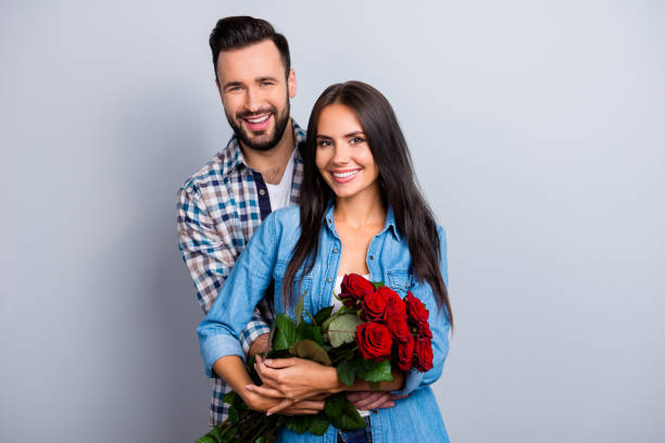 Portrait of cheerful lovely cute couple with beaming smiles hugging and looking at camera, woman hold a bouquet of red roses on 14 february, celebrating anniversary over grey background